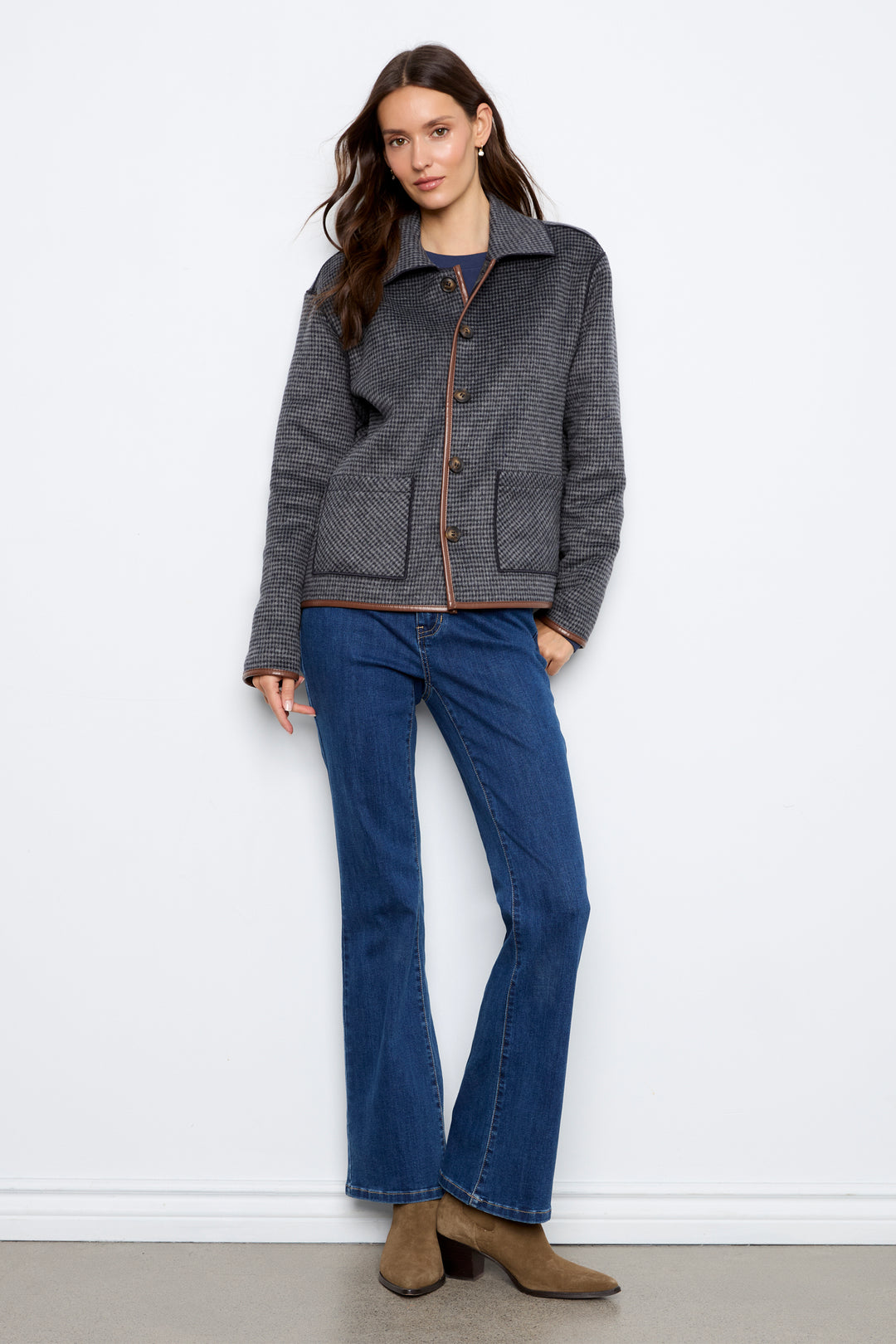 A woman modeling a dark gray textured jacket with brown buttons and brown trim, paired with blue jeans and brown ankle boots, standing against a white wall.