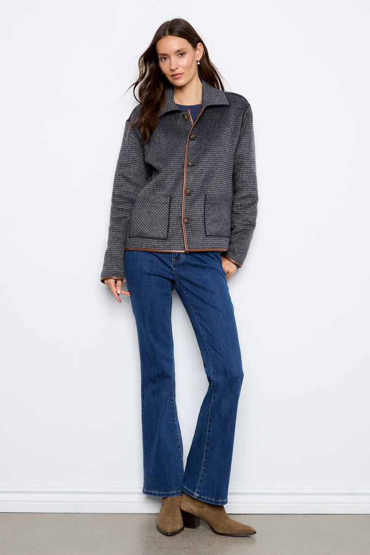 A woman modeling a dark gray textured jacket with brown buttons and brown trim, paired with blue jeans and brown ankle boots, standing against a white wall.