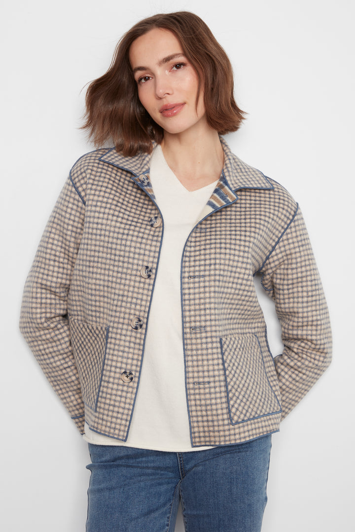 A woman wearing a beige and navy blue checkered jacket with blue piping, styled over a white top and blue jeans, against a plain light gray background.