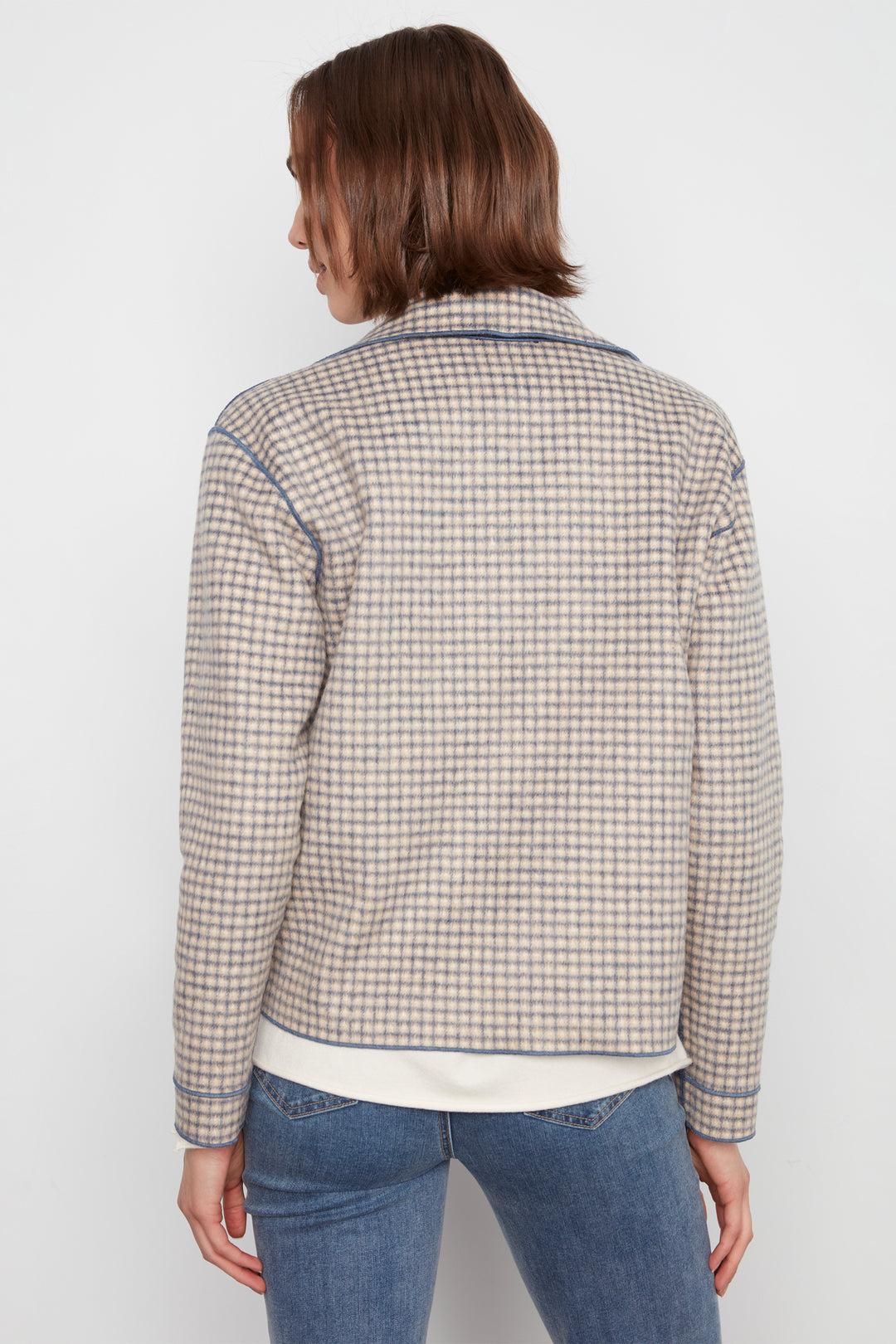 A woman shown from the back wearing a cream-colored plaid jacket with blue and brown stripes, paired with blue jeans, against a plain white background.