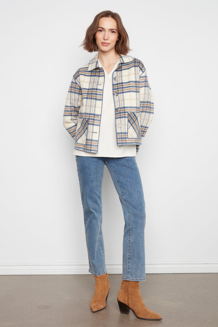 A woman stands against a plain white wall wearing a cream plaid jacket with blue and brown lines, a white t-shirt, blue jeans, and tan ankle boots.