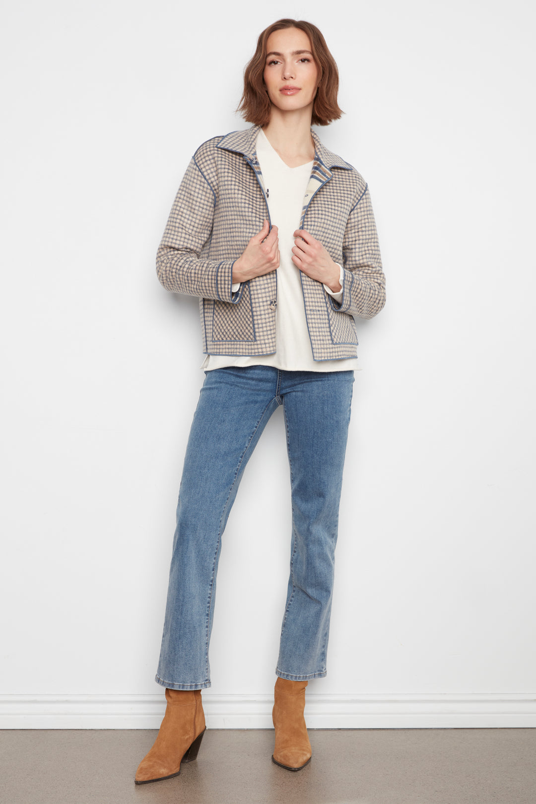 A woman stands against a white wall wearing a beige and gray patterned jacket over a white top, blue jeans, and tan ankle boots.