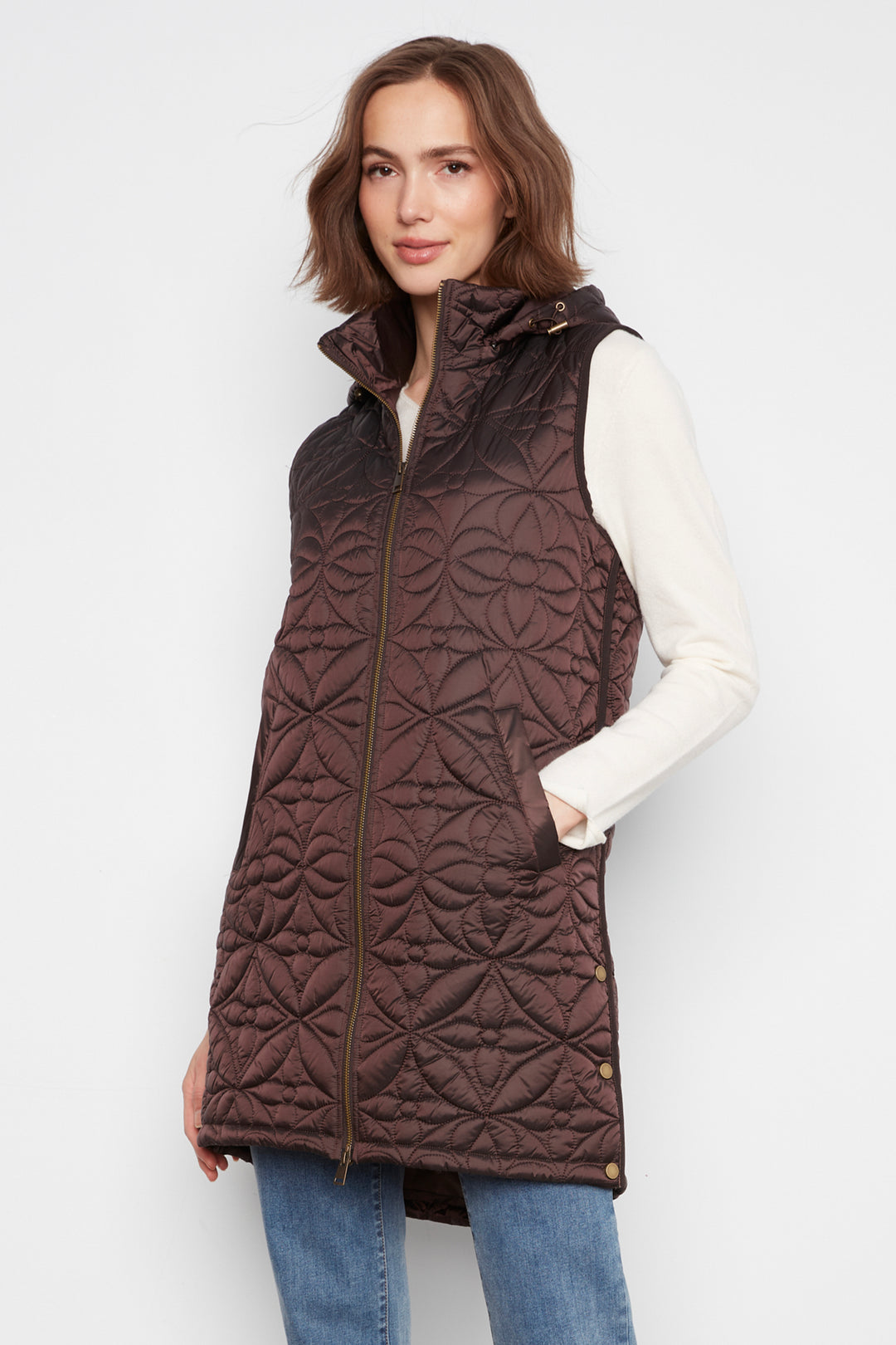 A woman wearing a dark brown quilted sleeveless puffer vest over a cream long-sleeve top and blue jeans, standing against a light gray studio background.