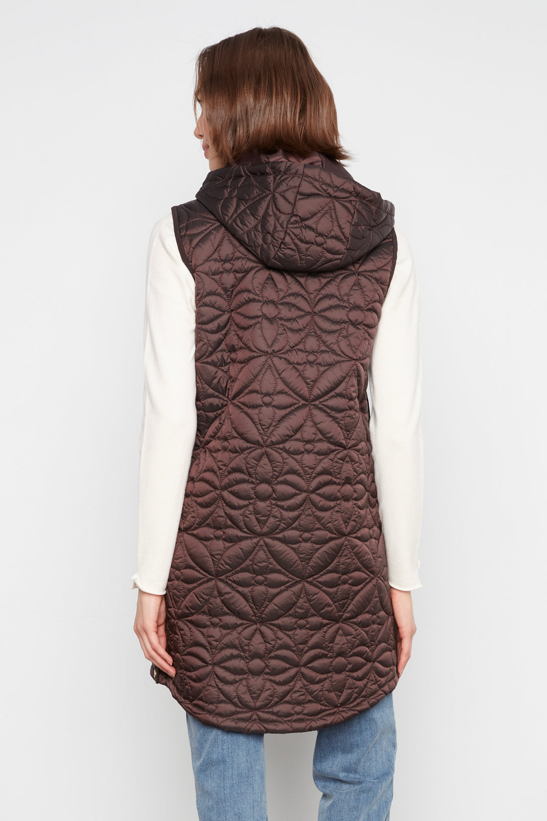 A woman shown from the back wearing a brown quilted hooded sleeveless vest over a cream long-sleeve top and blue jeans, against a plain light gray studio background.