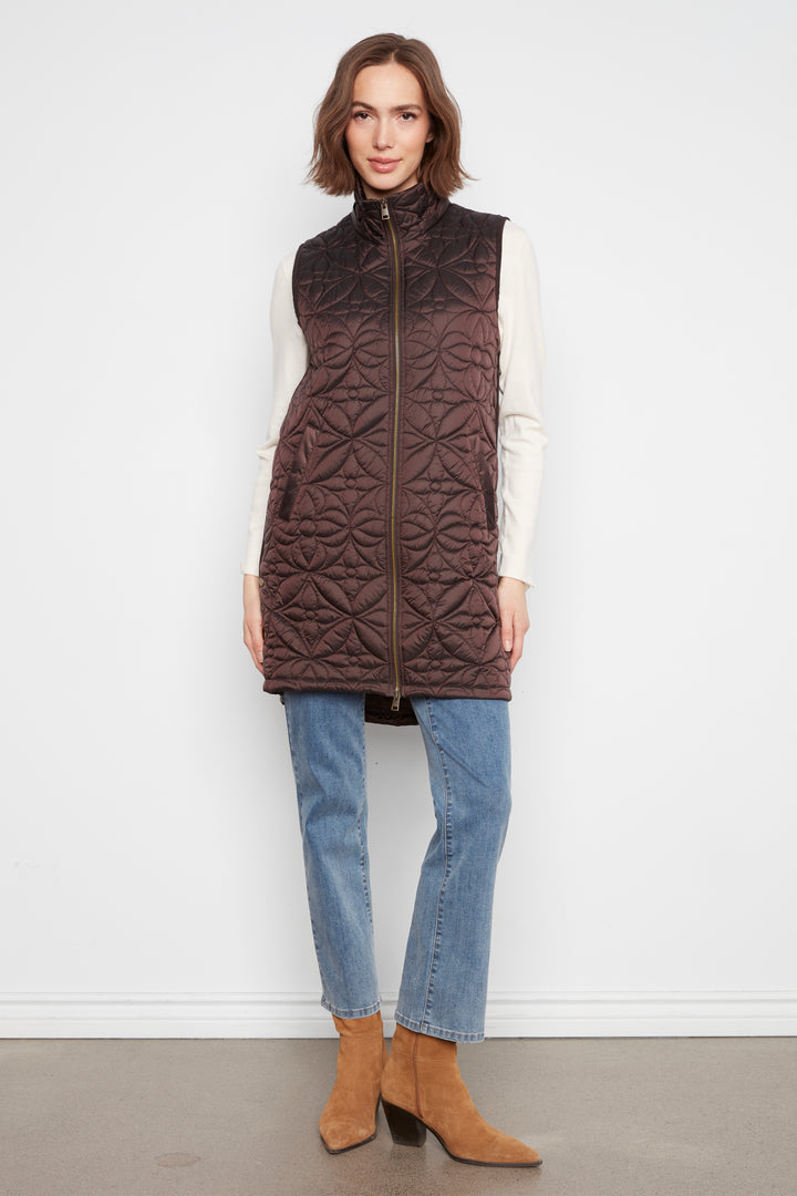 A woman stands against a plain white wall wearing a dark brown quilted sleeveless puffer vest over a cream long-sleeve top, blue jeans, and tan ankle boots.