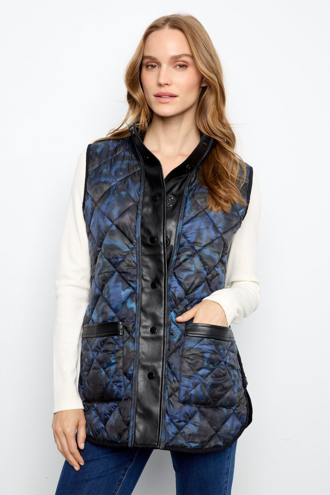 A woman modeling a sleeveless quilted puffer vest with a blue and black abstract pattern and black trim, worn over a white long-sleeve top and blue jeans against a plain white background.