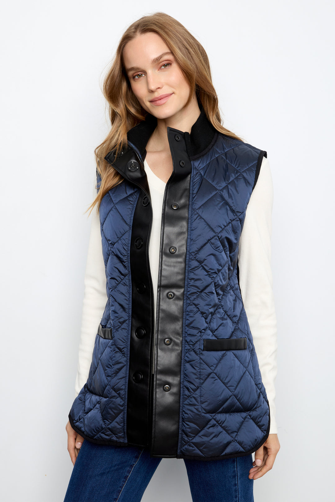 A woman wearing a navy blue quilted sleeveless vest with black faux-leather trim and black buttons, styled over a white long-sleeve top and blue jeans against a light gray studio background.