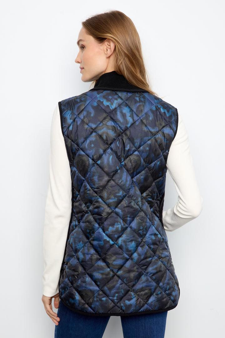 A woman shown from the back wearing a sleeveless diamond-quilted puffer vest over a white long-sleeve top and blue jeans. The vest features a blue and black abstract pattern.