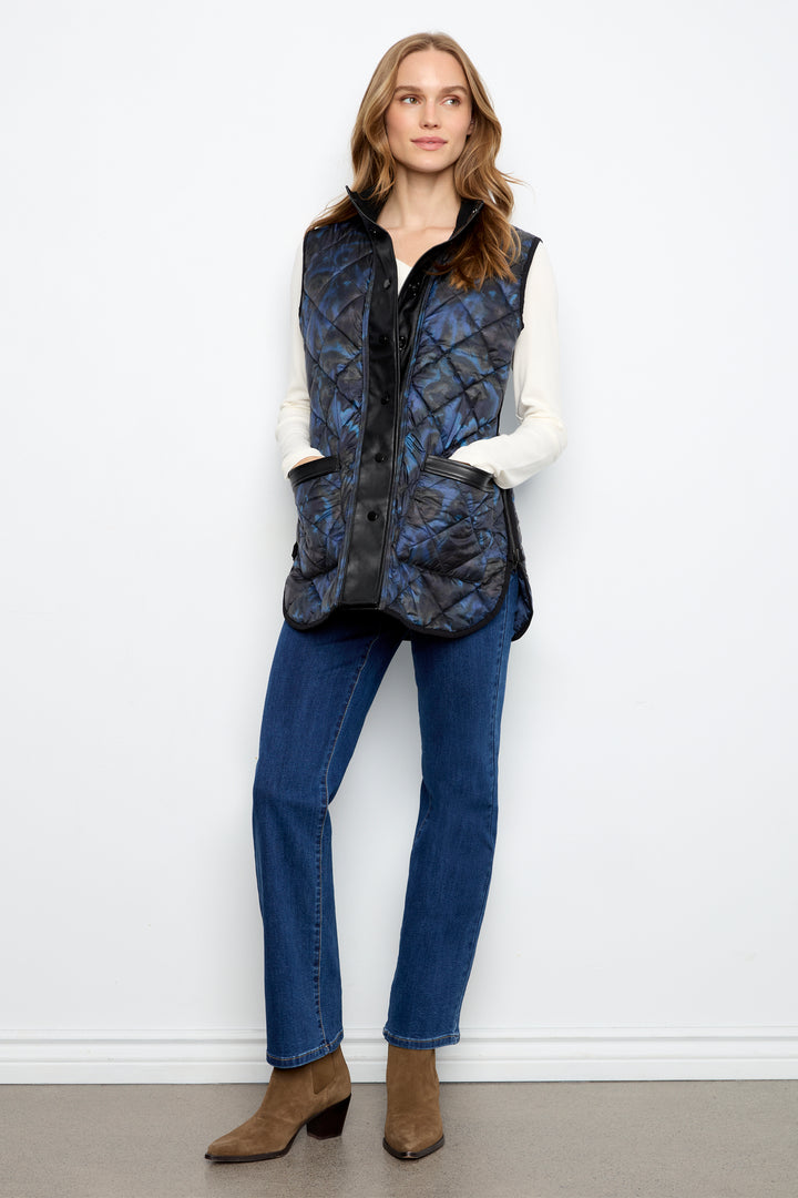 A woman modeling a sleeveless quilted vest with a blue and black abstract pattern, worn over a white long-sleeve shirt, blue jeans, and brown ankle boots against a plain white wall.