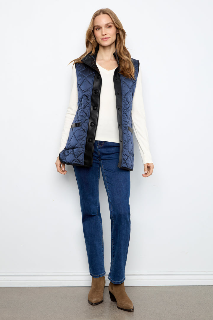 A woman stands against a white wall wearing a navy blue quilted sleeveless vest with black trim over a white long-sleeve top, blue jeans, and brown ankle boots.