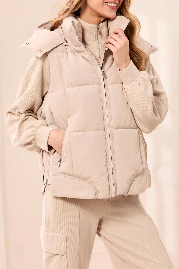 A woman wearing a light beige quilted puffer vest over a matching beige long-sleeve top and beige pants, against a soft beige studio background.