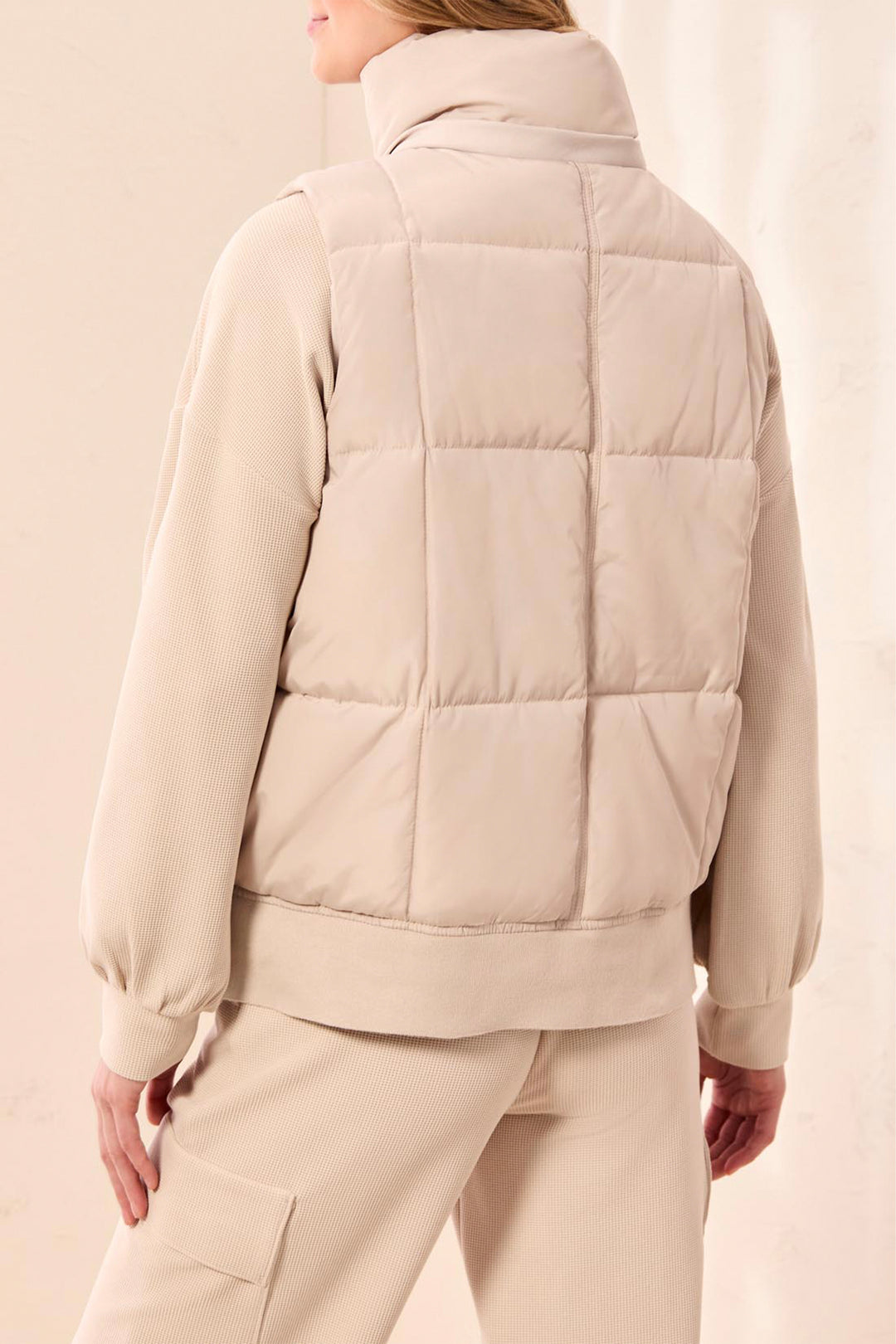 A woman shown from the back wearing a light beige quilted puffer vest over a matching beige long-sleeve top and beige pants, against a soft beige studio background.
