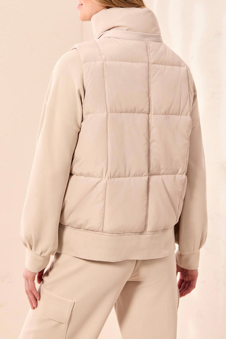 A woman shown from the back wearing a light beige quilted puffer vest over a matching beige long-sleeve top and beige pants, against a soft beige studio background.