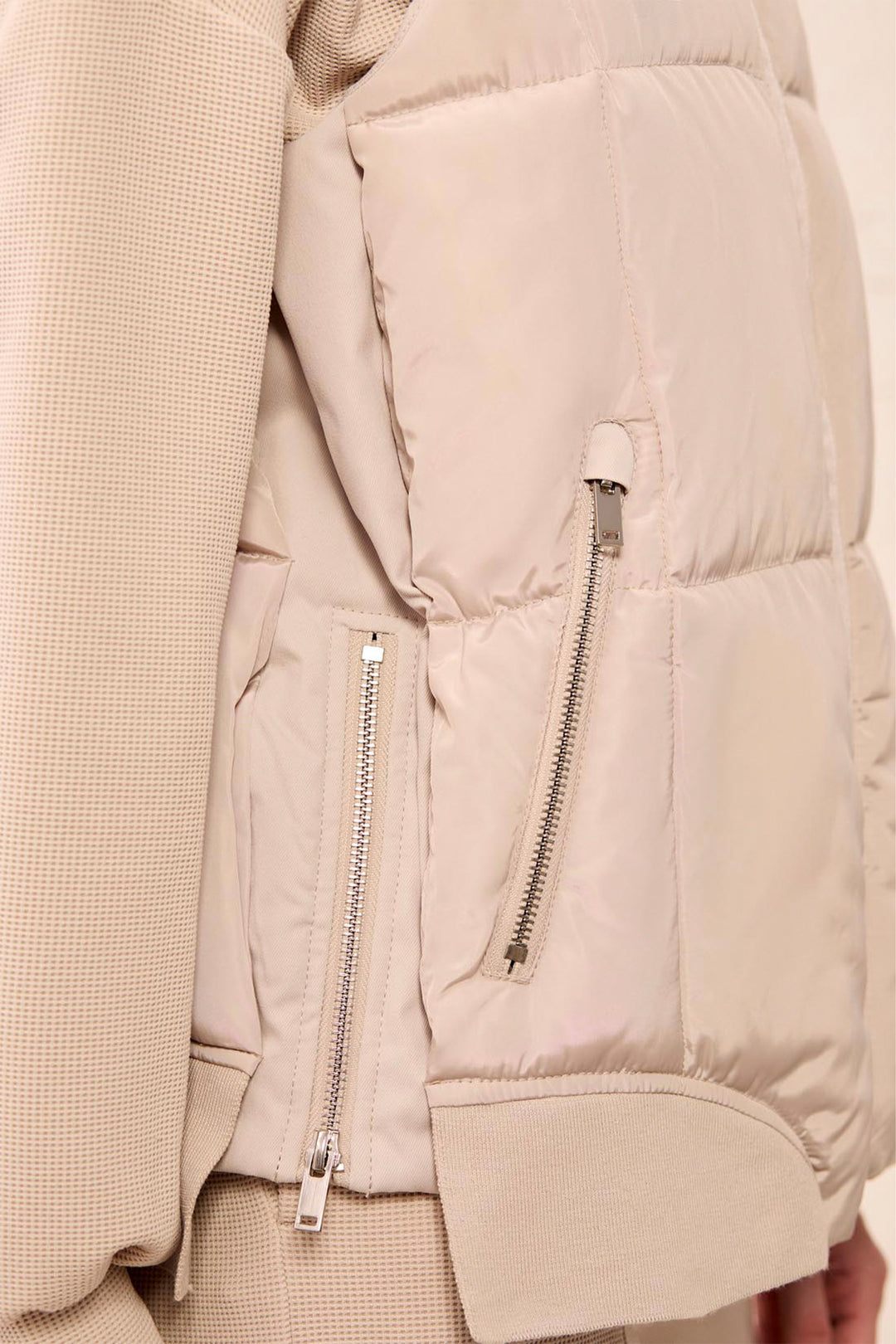 Close-up of a light beige puffer vest with silver zippers, shown on a model wearing a matching textured knit sweater.