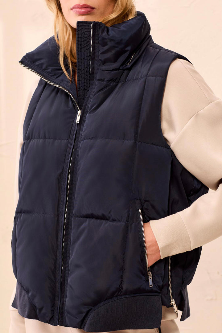 A close-up of a model wearing a navy blue puffer vest over a cream-colored top. The vest features a high collar, a front zipper, and zippered side pockets.