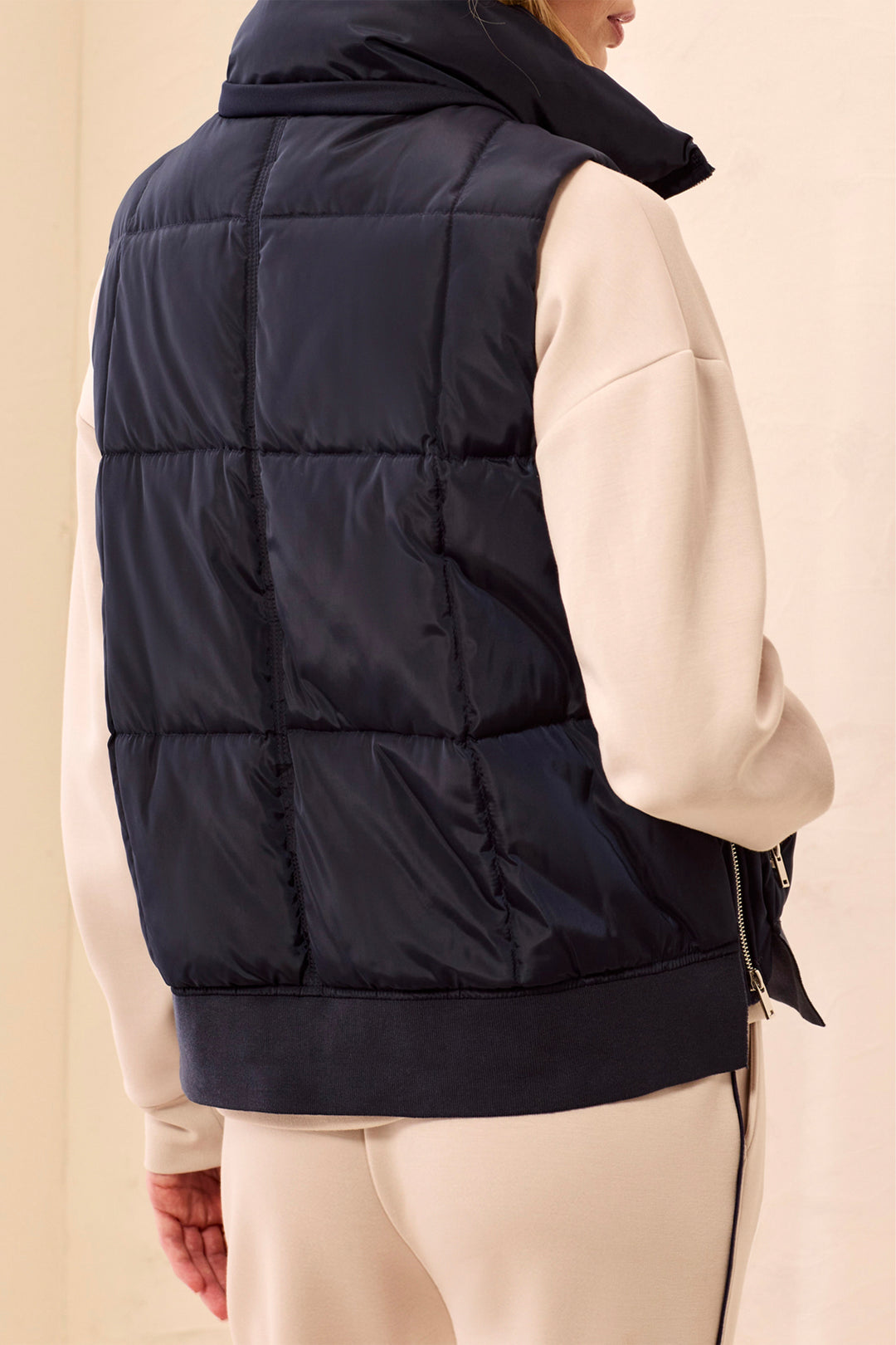 A person shown from the back wearing a dark navy puffer vest over a cream-colored sweatshirt and cream pants, against a plain beige background.