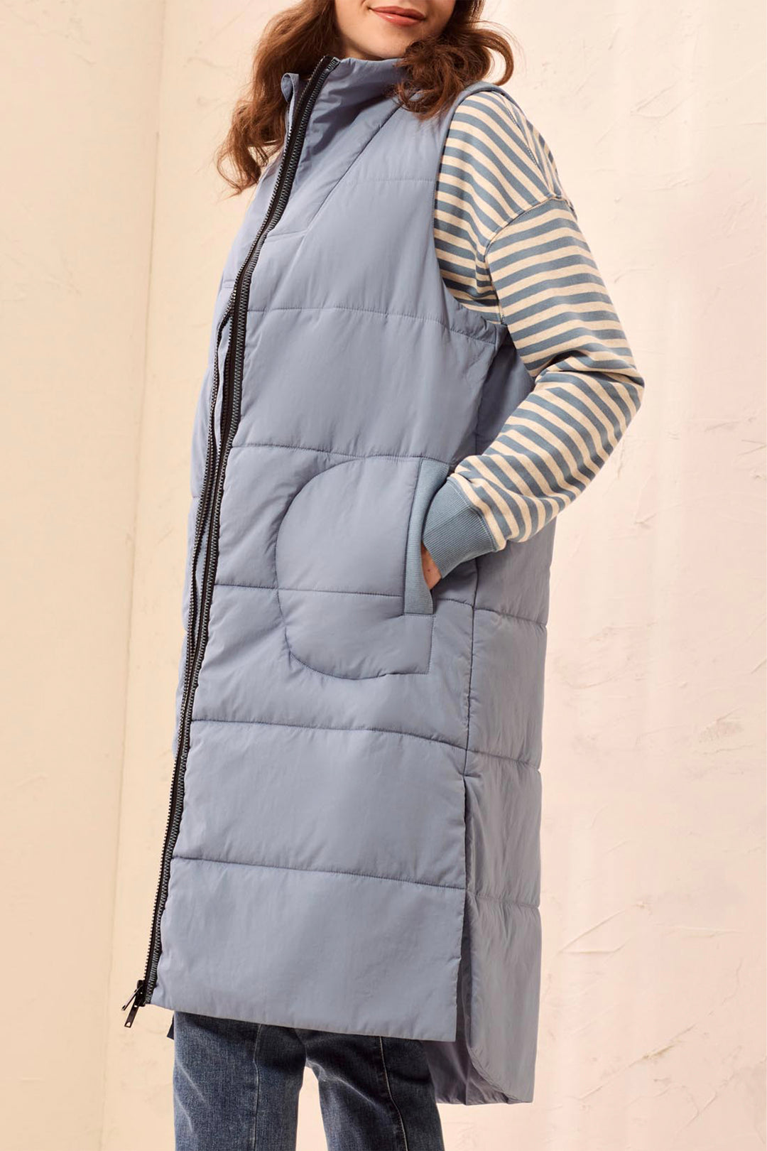 A woman wearing a long, slate blue-gray quilted puffer vest over a beige and blue striped long-sleeve top, paired with blue jeans, set against a light beige textured wall.