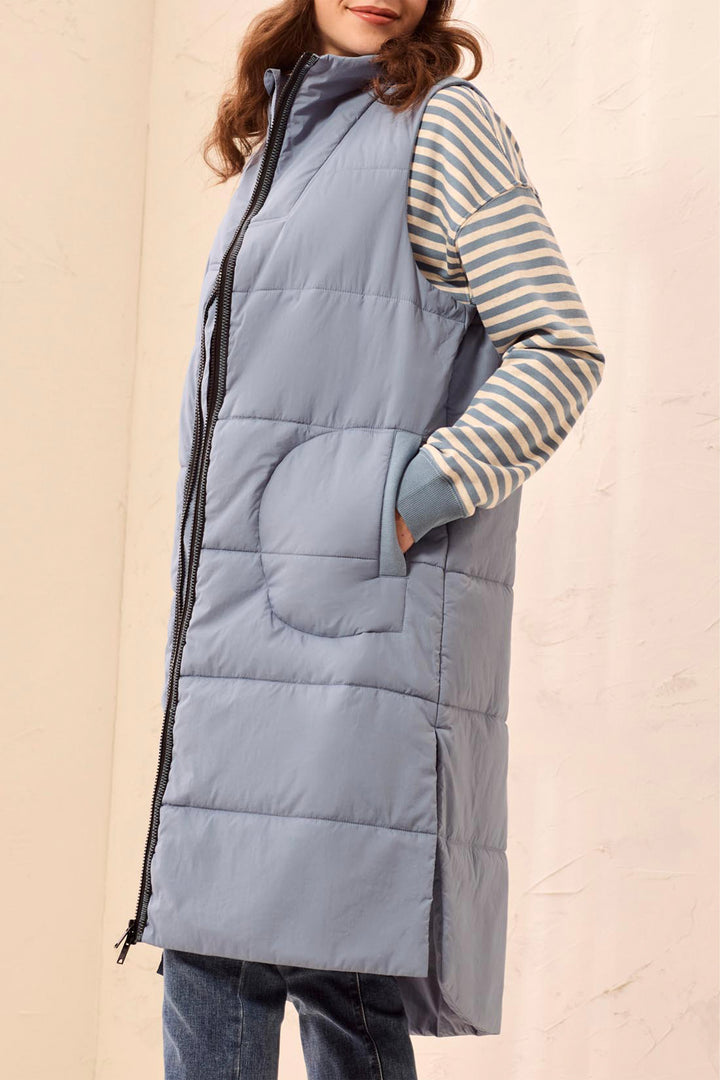 A woman wearing a long, slate blue-gray quilted puffer vest over a beige and blue striped long-sleeve top, paired with blue jeans, set against a light beige textured wall.