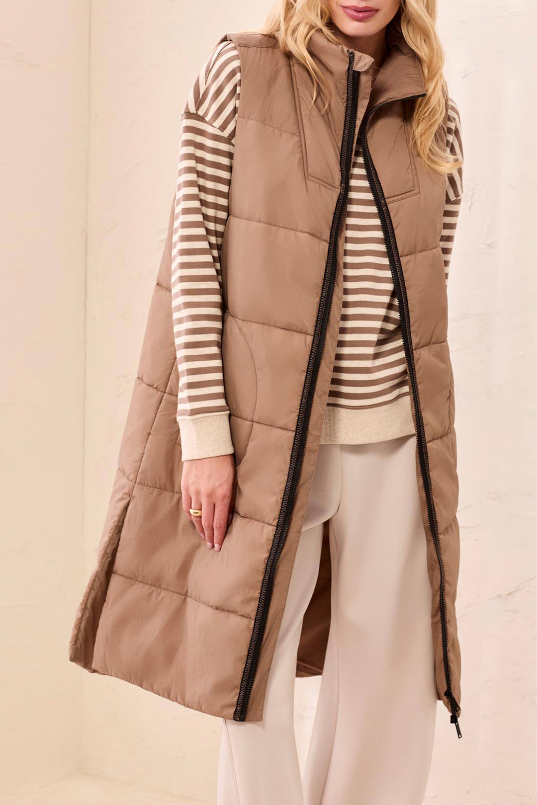 Woman wearing a long brown puffer vest over a striped shirt with a beige background