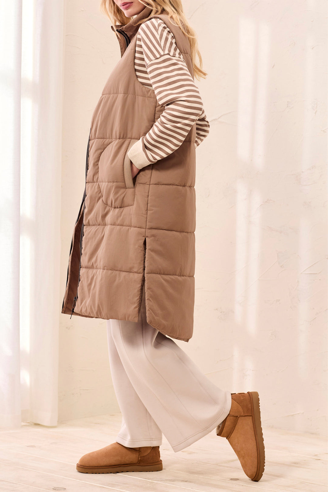 Person wearing a brown puffer vest over a striped shirt with white pants and brown shoes, standing against a light curtain background.