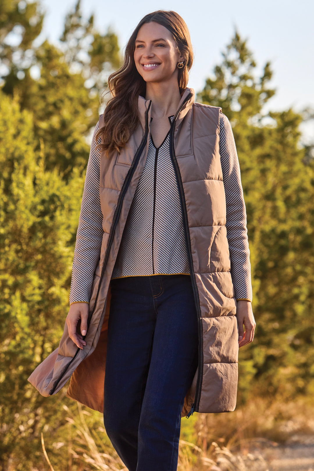 A woman wearing a long taupe quilted puffer vest over a patterned knit sweater and dark blue jeans, standing outdoors in front of a soft-focus background of green trees and dry grass.