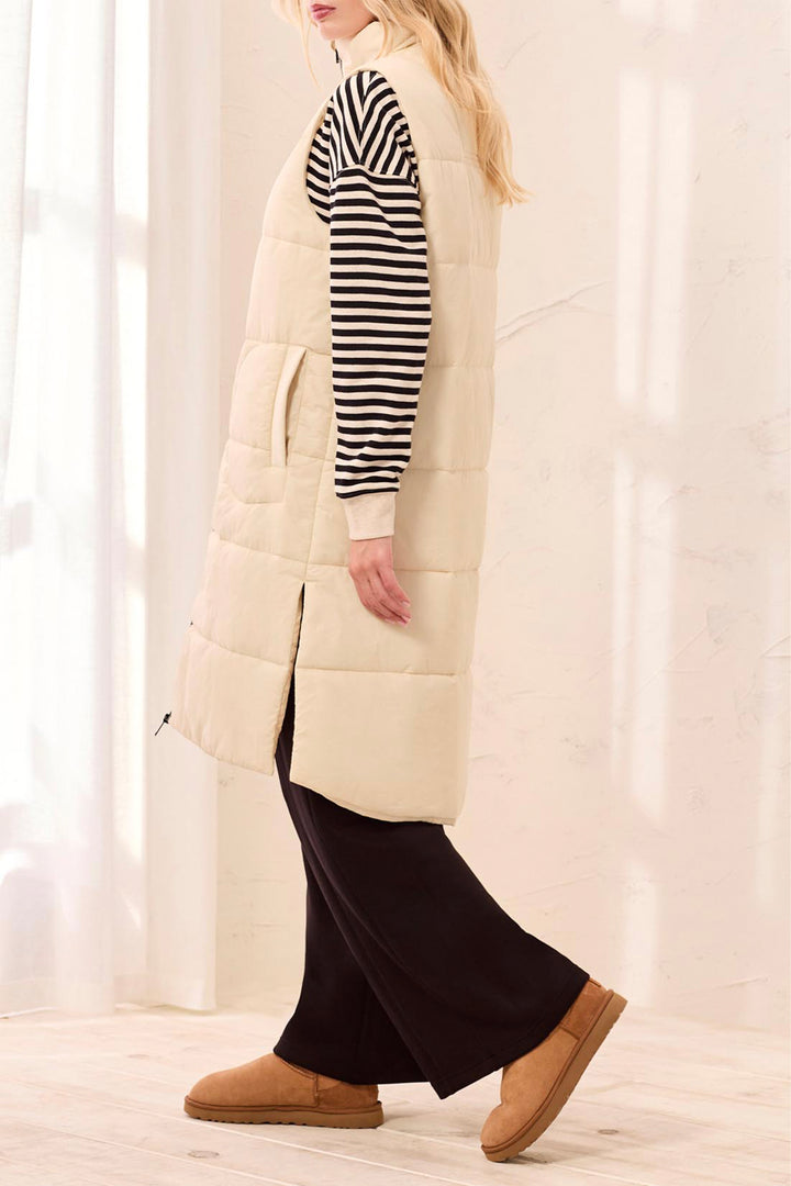 A woman wearing a long cream quilted puffer vest over a black and white striped long-sleeve top, black wide-leg pants, and tan slip-on shoes, standing in a bright indoor setting with soft window light.