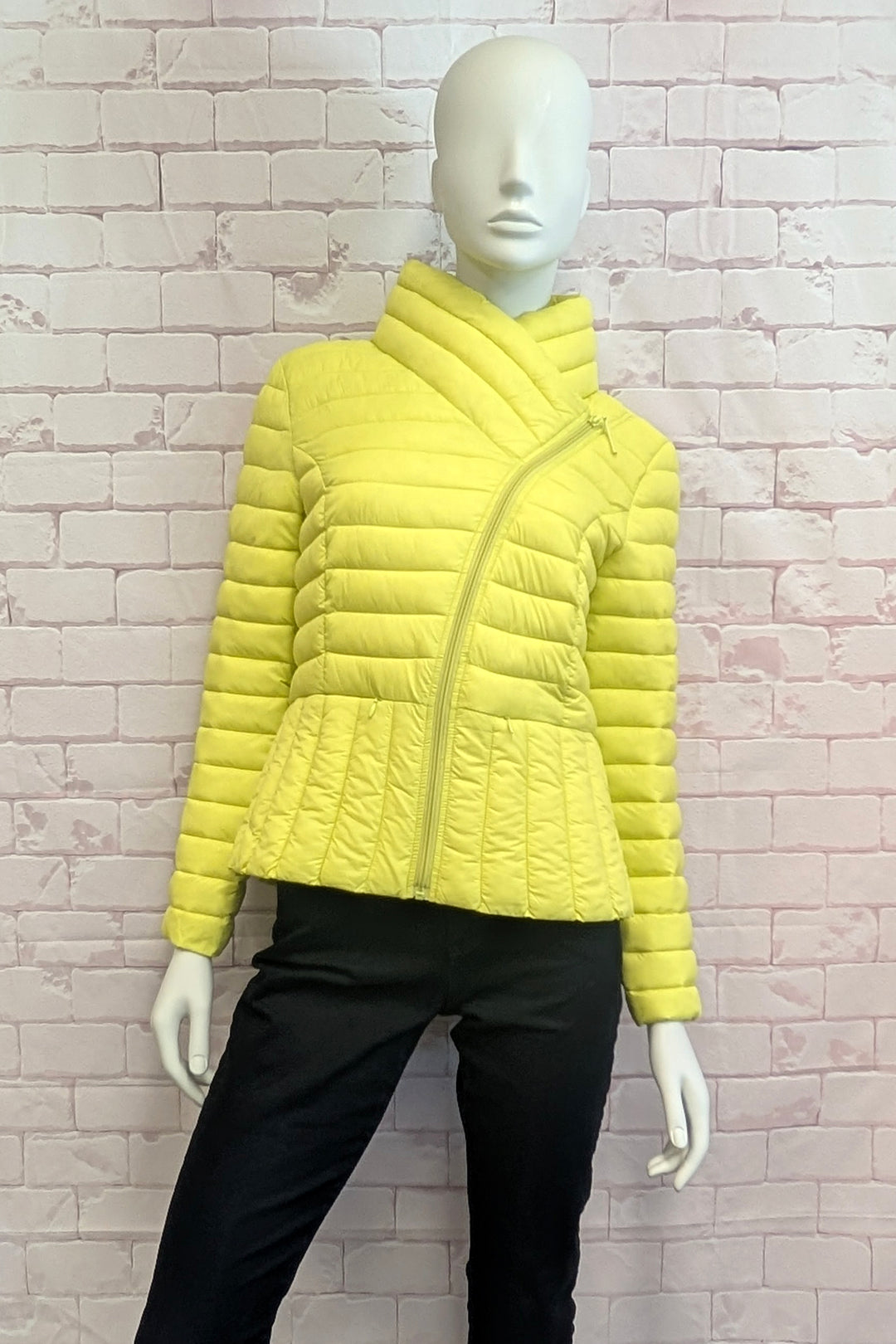A bright yellow quilted puffer jacket with an asymmetrical zipper and high stand collar, displayed on a white mannequin torso against a light pink brick wall.