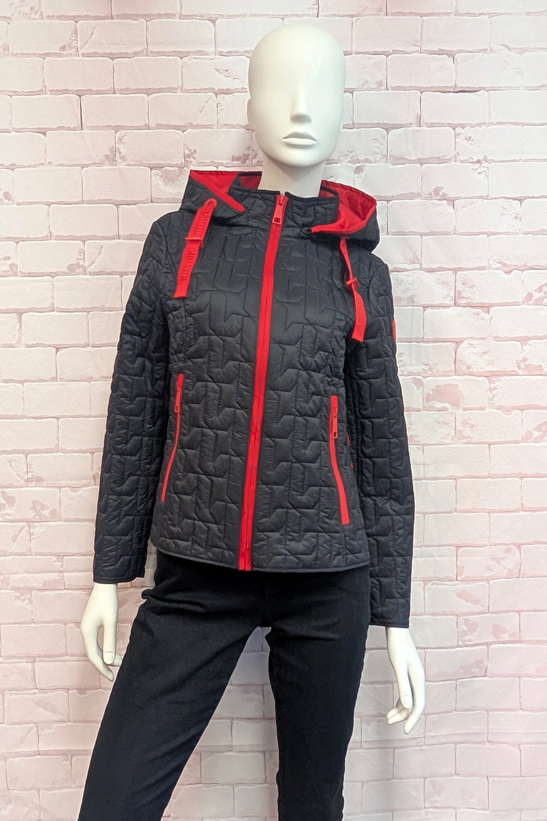 A black quilted hooded jacket with bright red trim and zipper details, displayed on a white mannequin against a light pink brick wall.