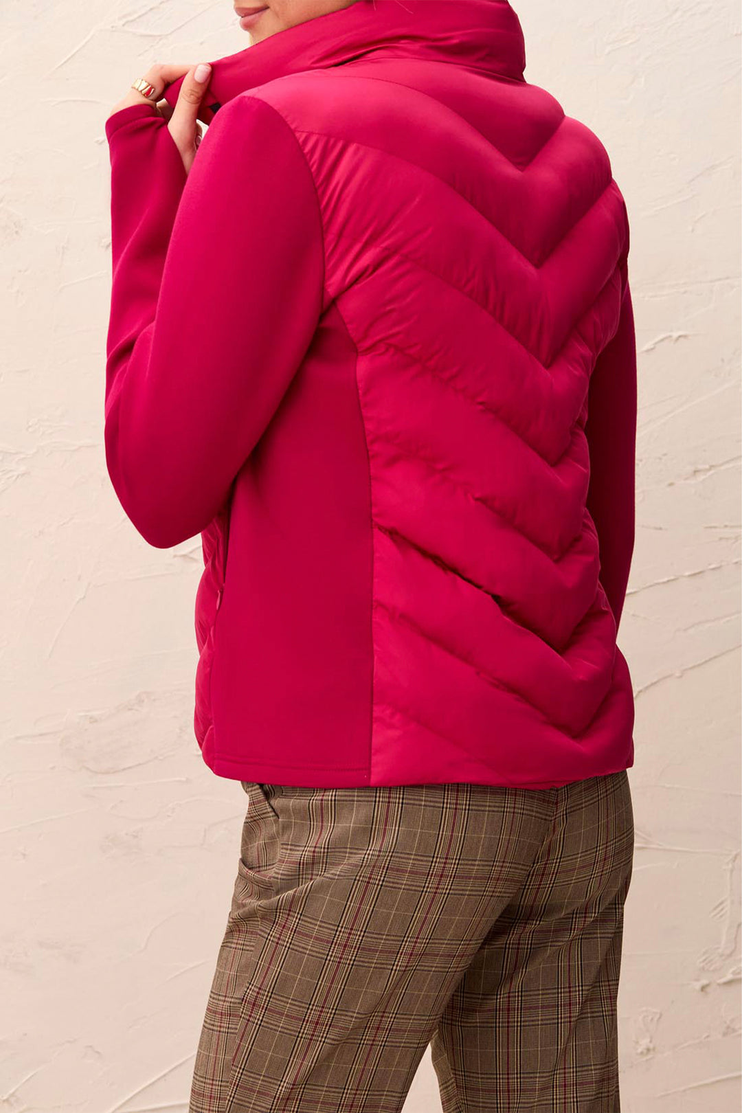 A person wearing a bright pink quilted puffer jacket with a high collar, shown from the back against a textured off-white wall.