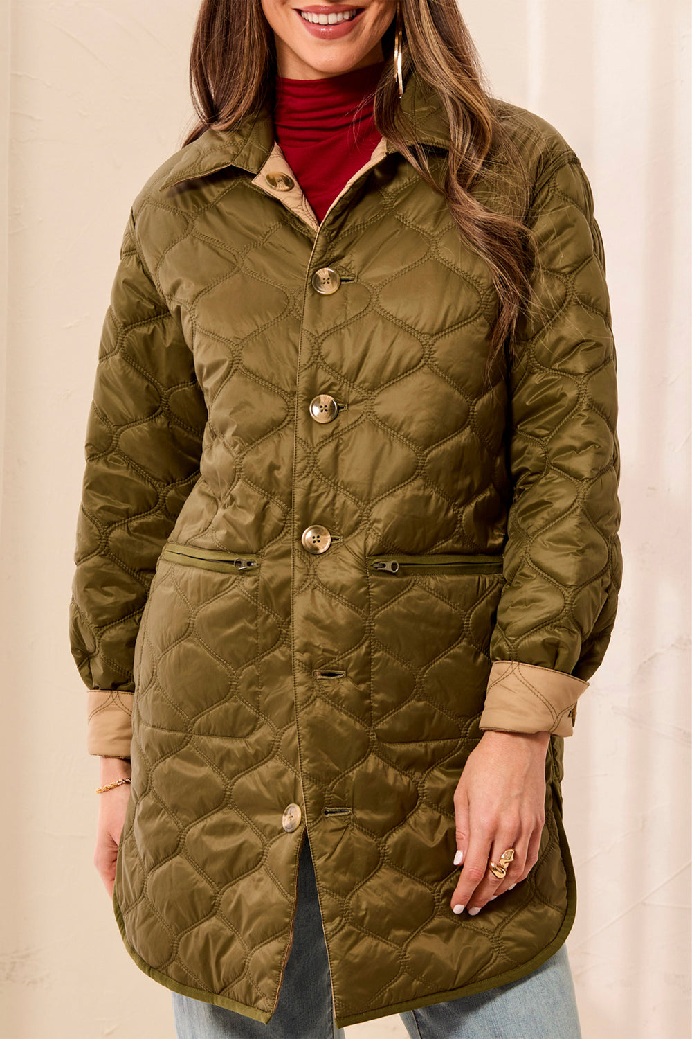 A woman wearing a quilted olive green coat with tan lining and gold-tone buttons, layered over a red turtleneck and light blue jeans, against a neutral cream background.