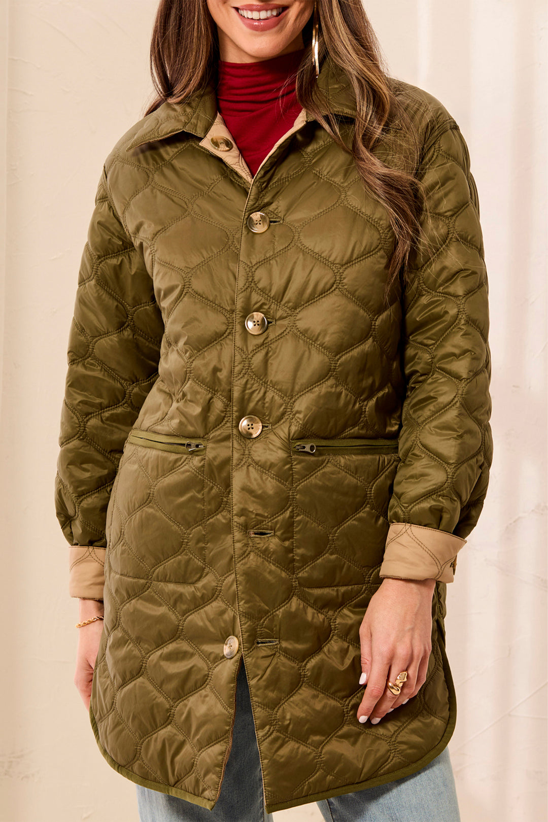 A woman wearing a quilted olive green coat with tan lining and gold-tone buttons, layered over a red turtleneck and light blue jeans, against a neutral cream background.