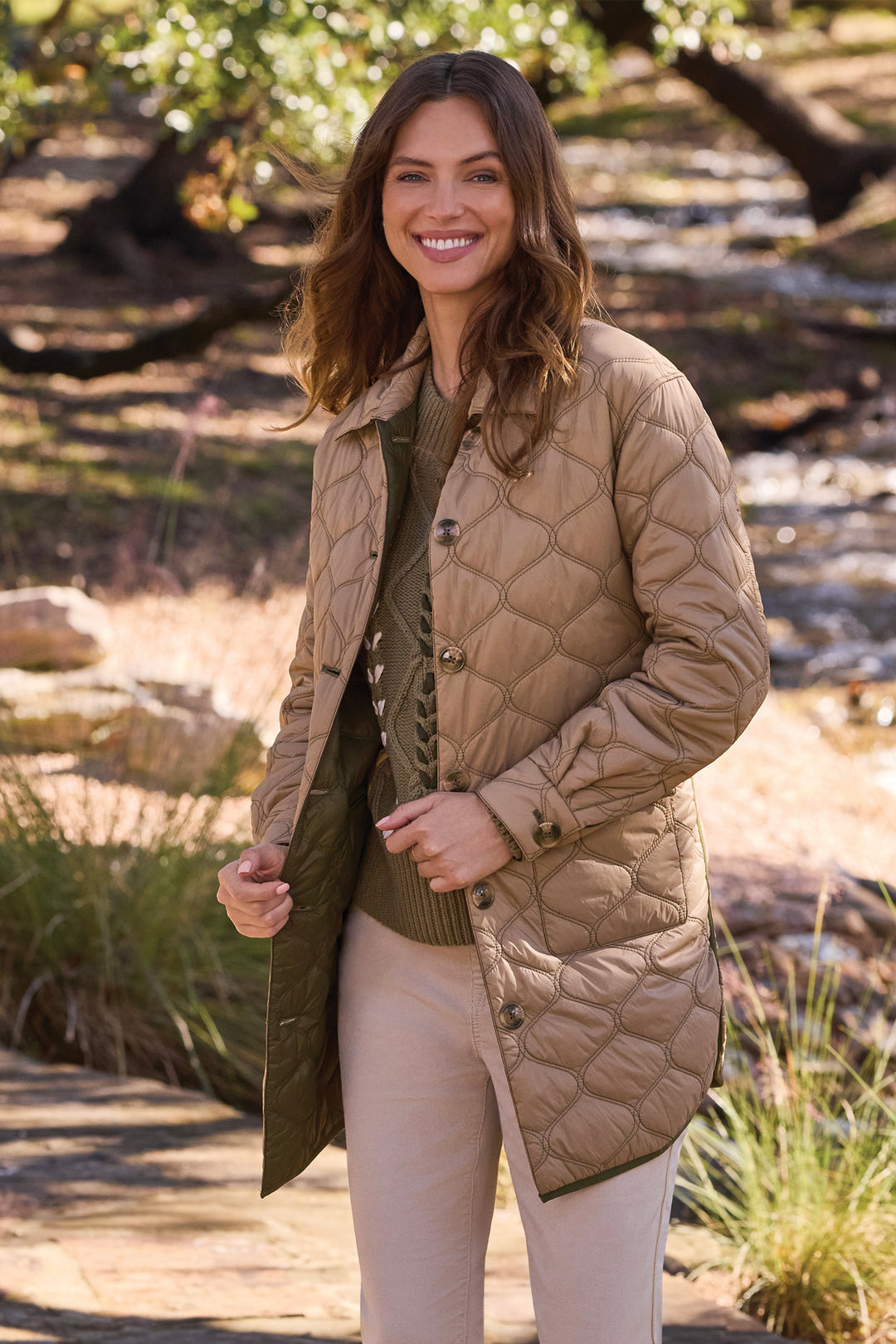 A woman wearing a tan quilted mid-length jacket over an olive green sweater and beige pants, standing outdoors in a park-like setting with trees and a stone path.
