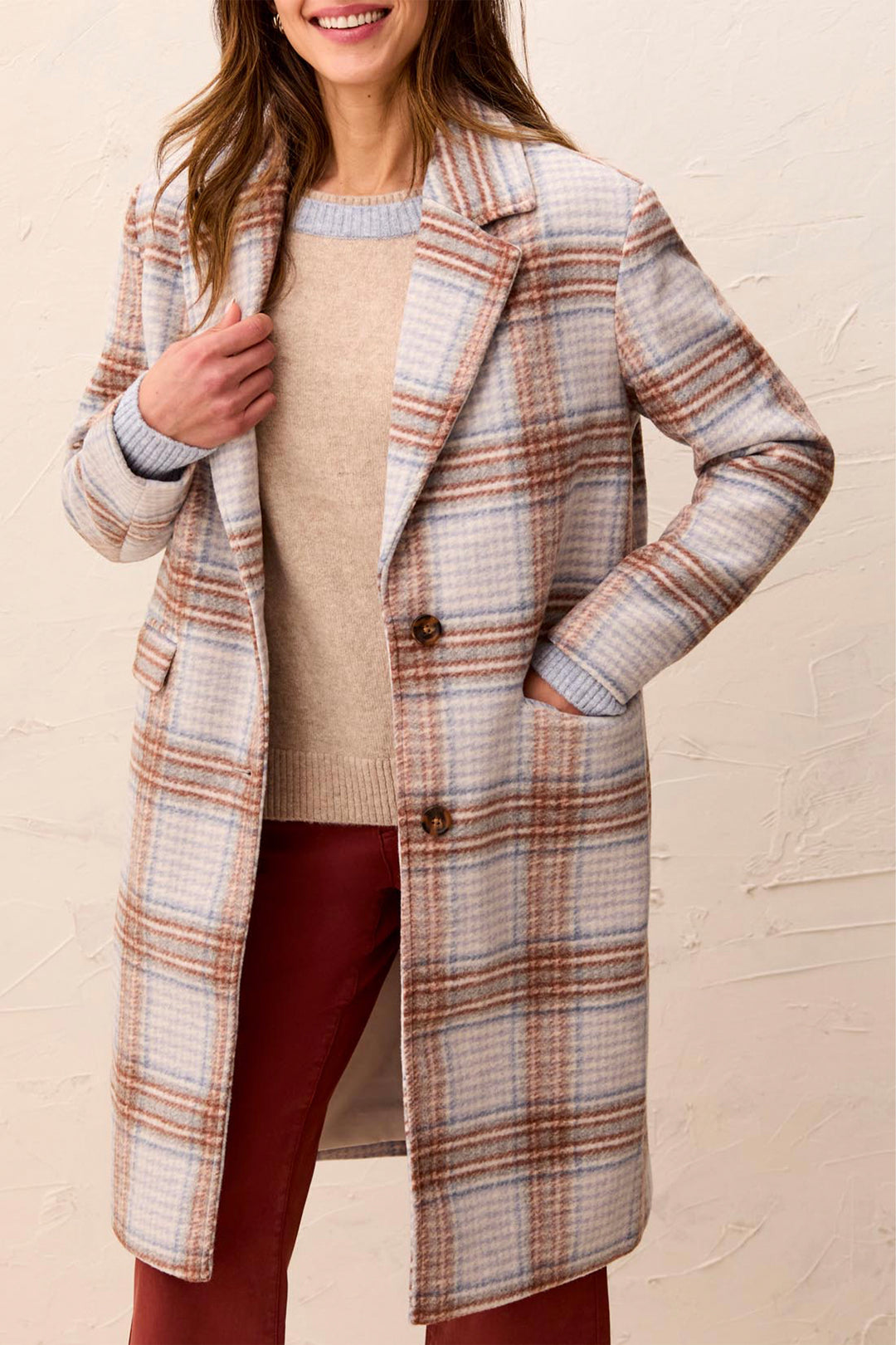 A woman wearing a long plaid coat over a beige sweater and burgundy pants, standing against a textured beige wall.