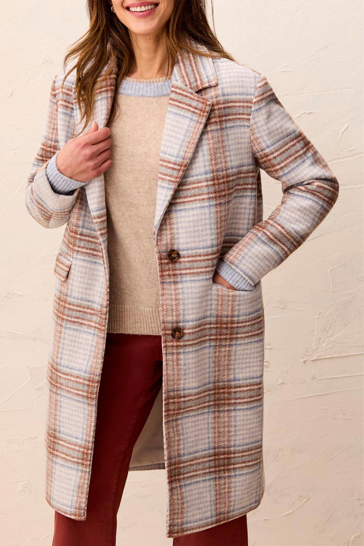 A woman wearing a long plaid coat over a beige sweater and burgundy pants, standing against a textured beige wall.