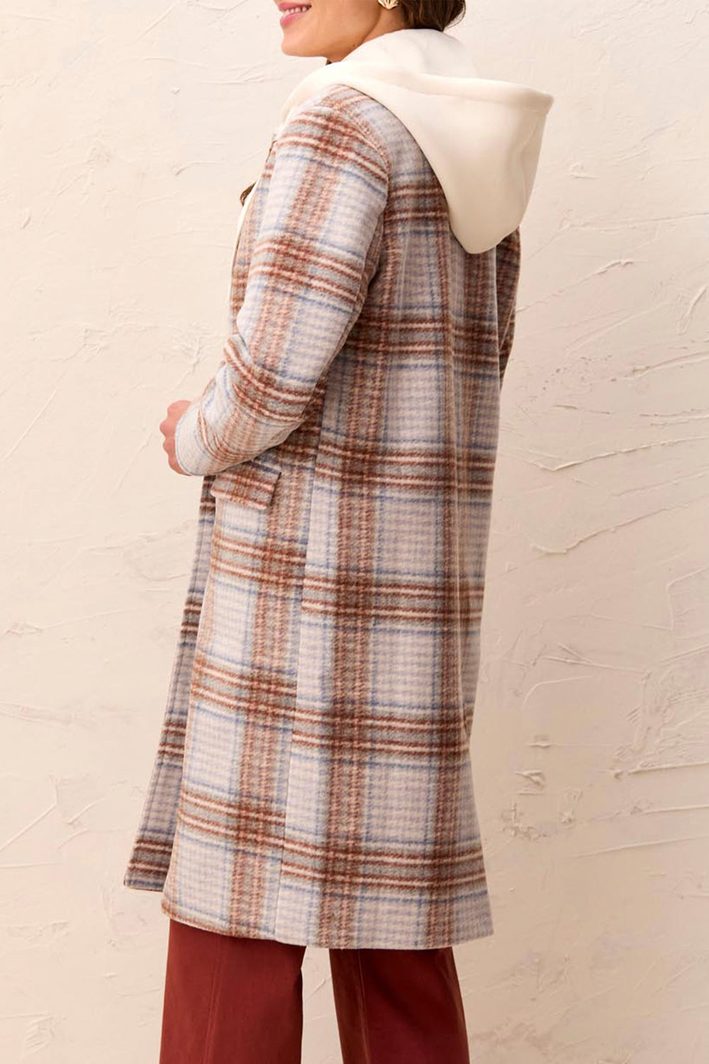 A woman wearing a long plaid coat shown from the back over a beige sweater and burgundy pants, standing against a textured beige wall.