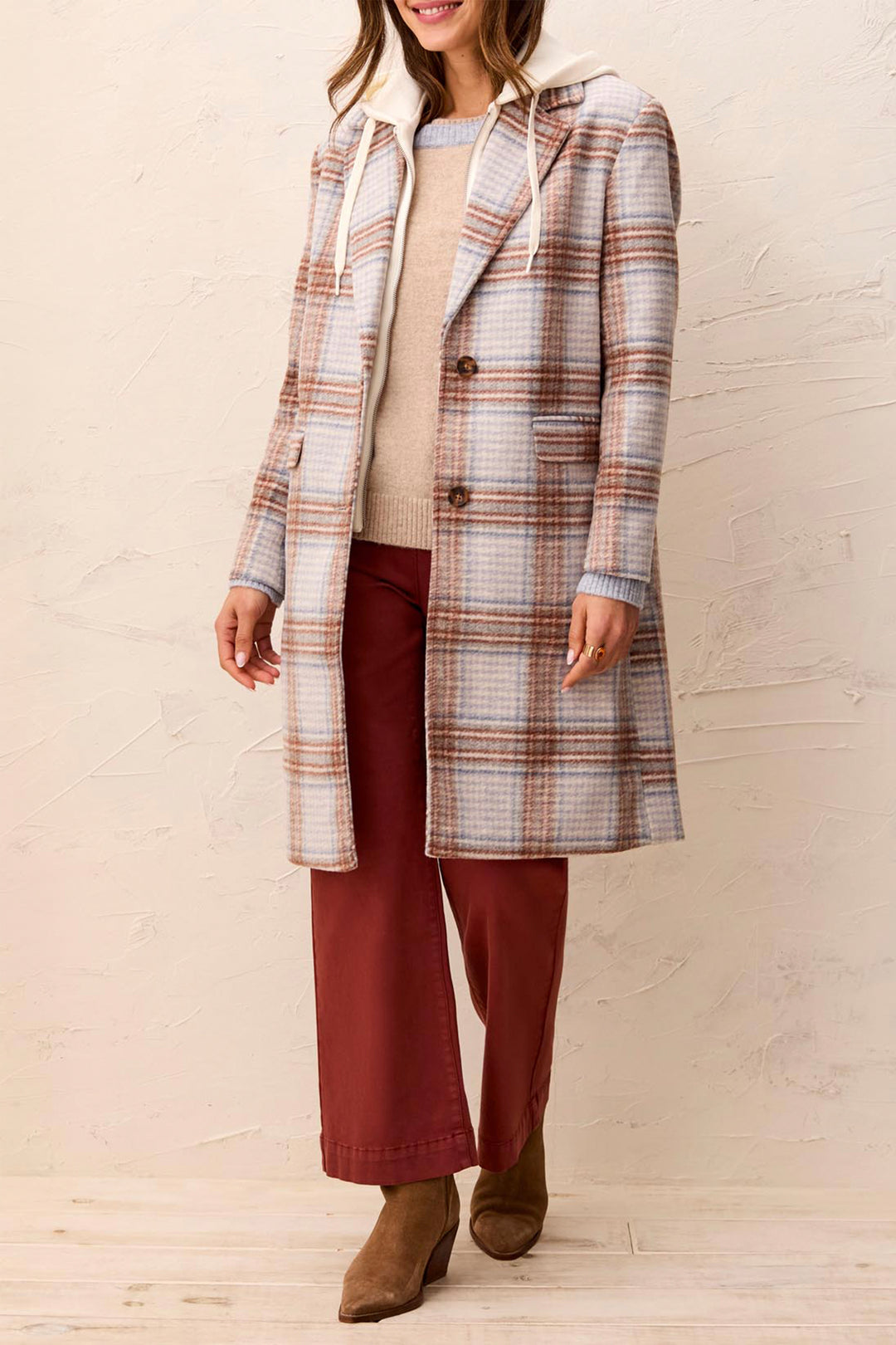 A woman wearing a long plaid coat over a beige sweater and burgundy pants, standing against a textured beige wall.