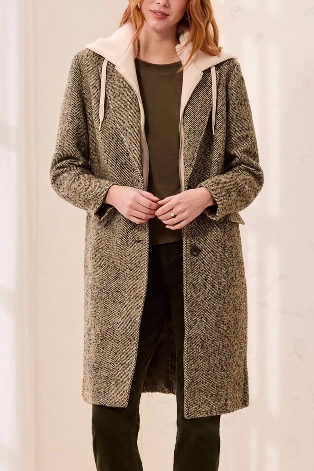 A woman wearing a long, textured herringbone coat over a cream-colored hooded sweatshirt, paired with dark pants, against a plain light background.