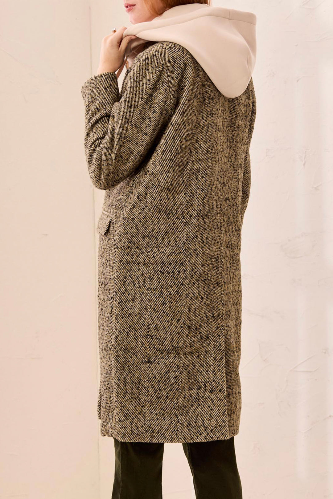 A woman wearing a long, textured herringbone coat over a cream-colored hooded sweatshirt, paired with dark pants, shown from a side-back angle against a plain light background.