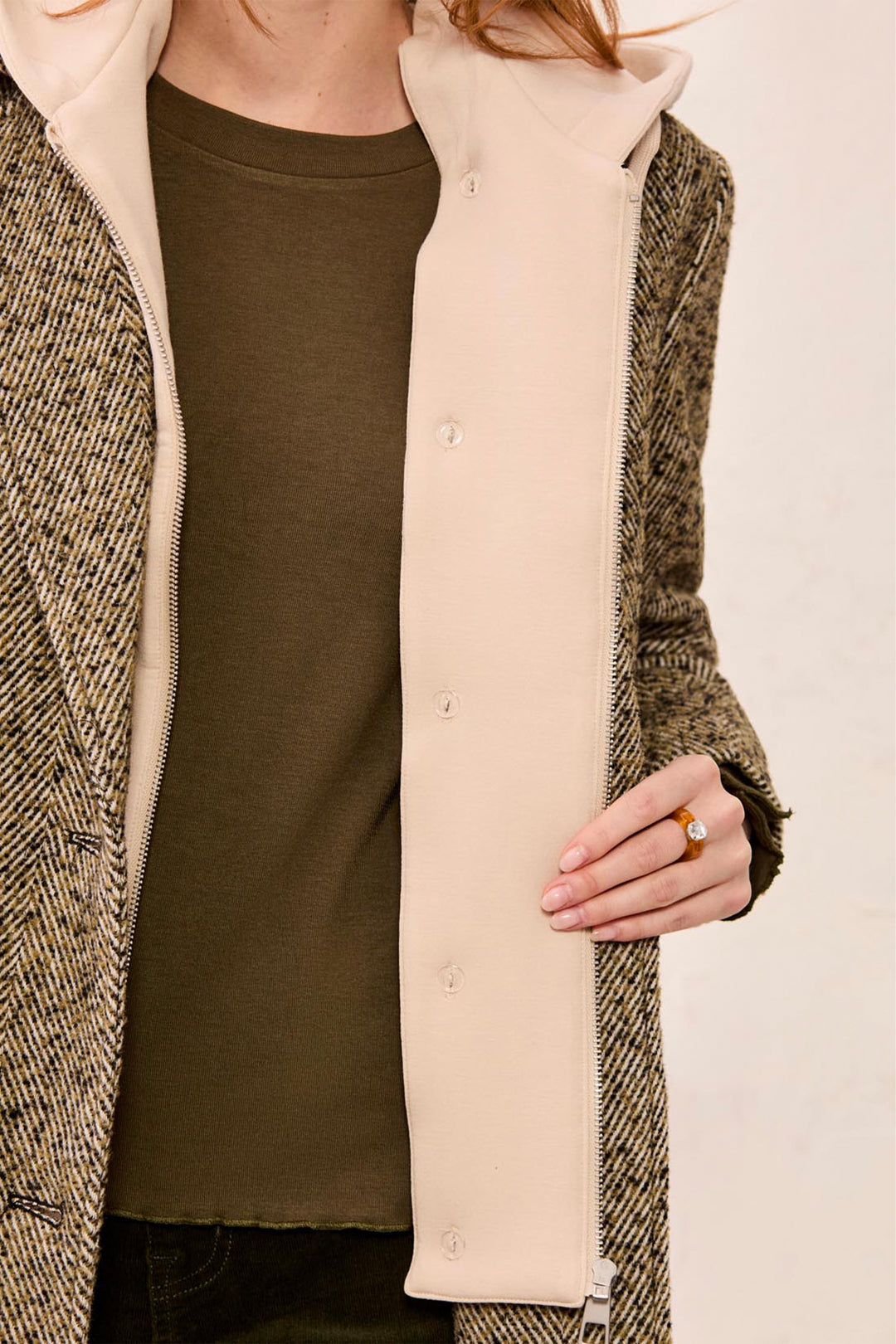 A close-up of a person wearing a beige fleece zippered hooded vest over a dark olive green top, paired with a brown and beige herringbone coat.