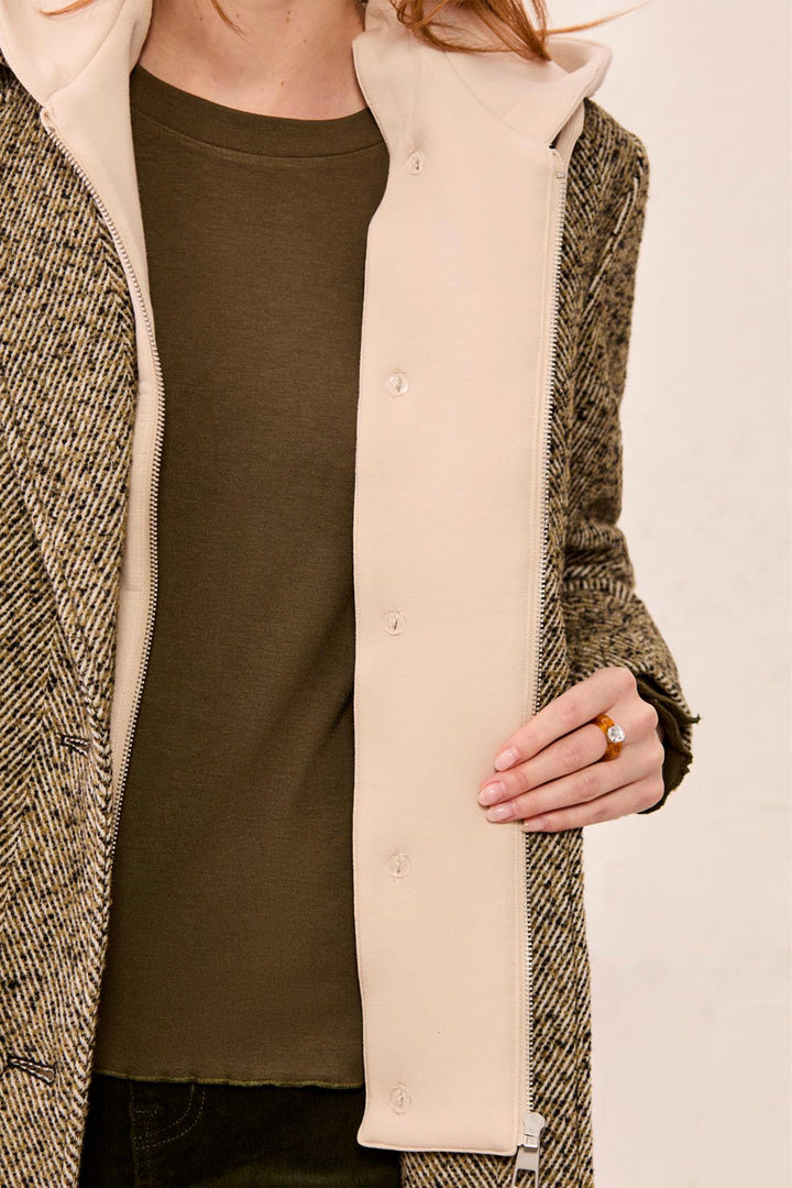 A close-up of a person wearing a beige fleece zippered hooded vest over a dark olive green top, paired with a brown and beige herringbone coat.