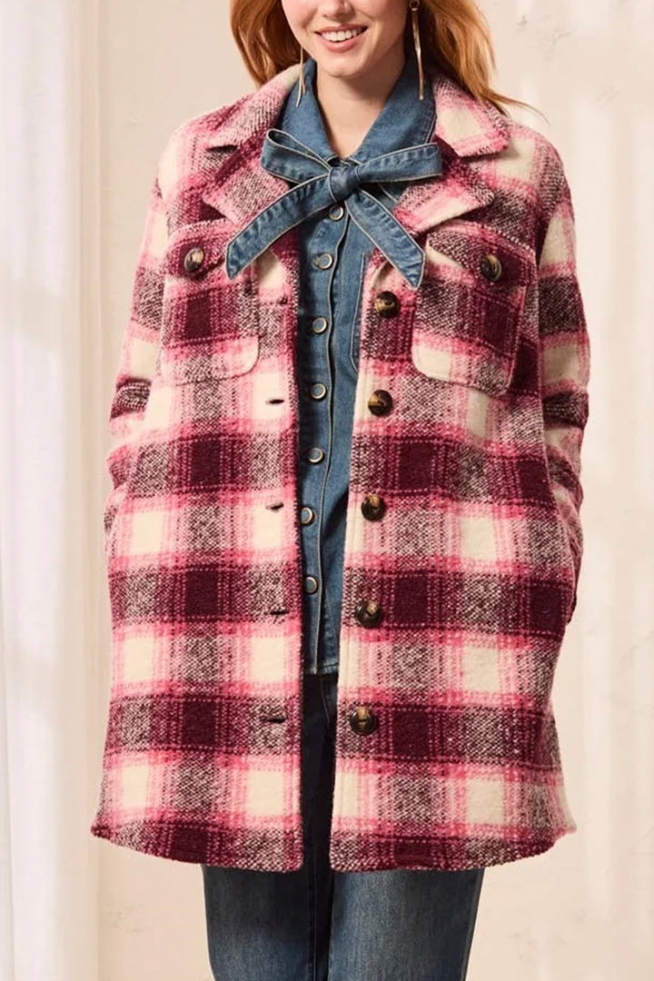 A woman wearing a mid-length plaid coat in pink, burgundy, and cream, paired with blue jeans, angle against a light beige wall.