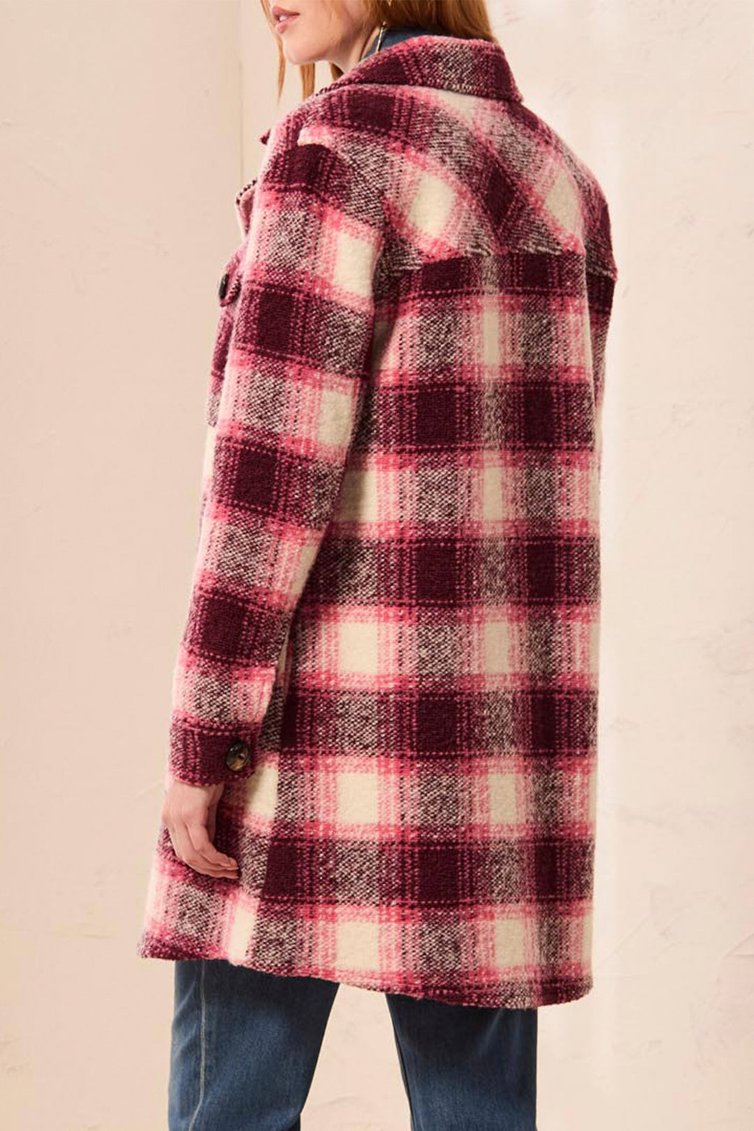 A woman wearing a mid-length plaid coat in pink, burgundy, and cream, paired with blue jeans, shown from a side-back angle against a light beige wall.