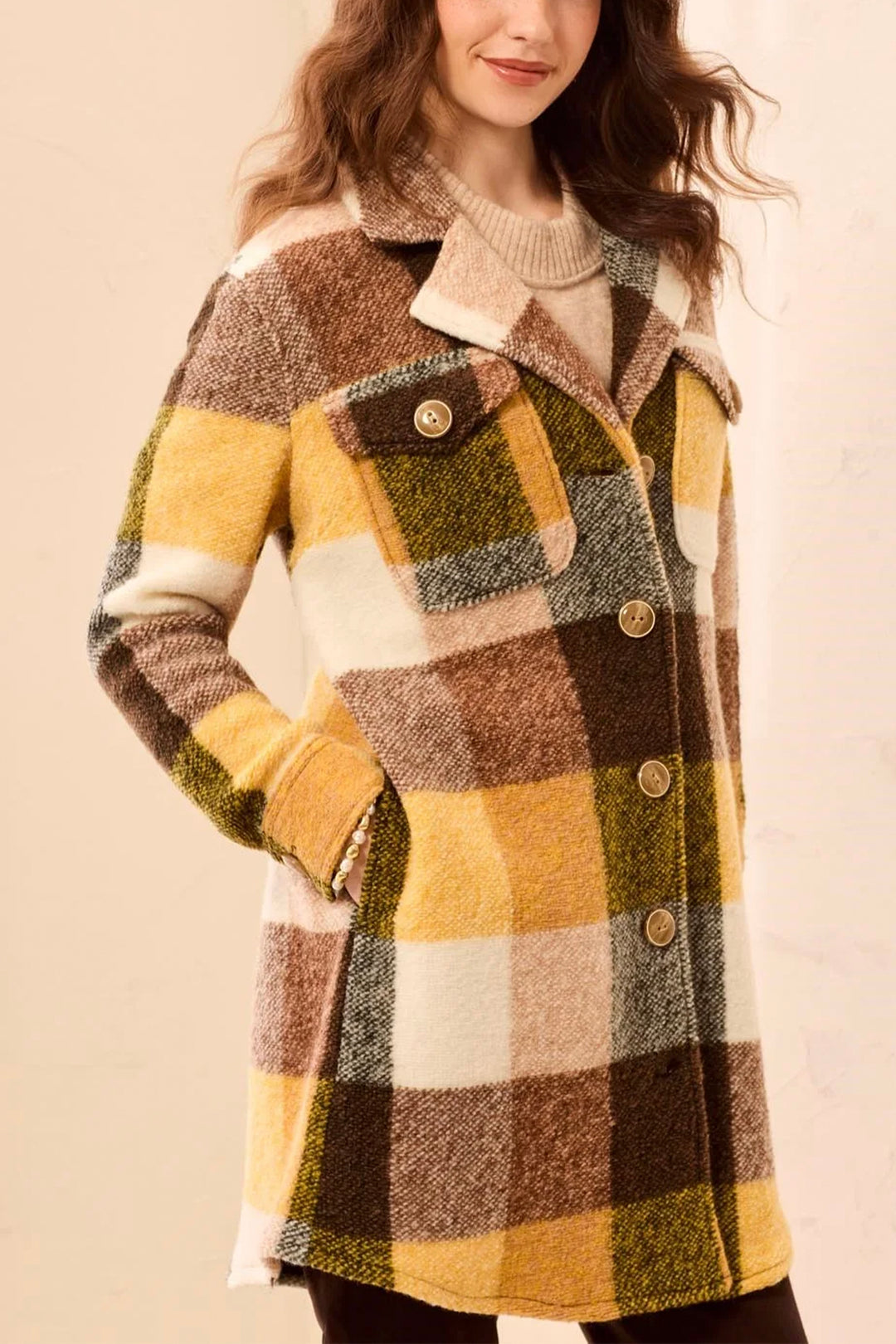 A woman wearing a long plaid coat in mustard yellow, brown, cream, and olive green, paired with a beige knit sweater,  against a soft beige background.