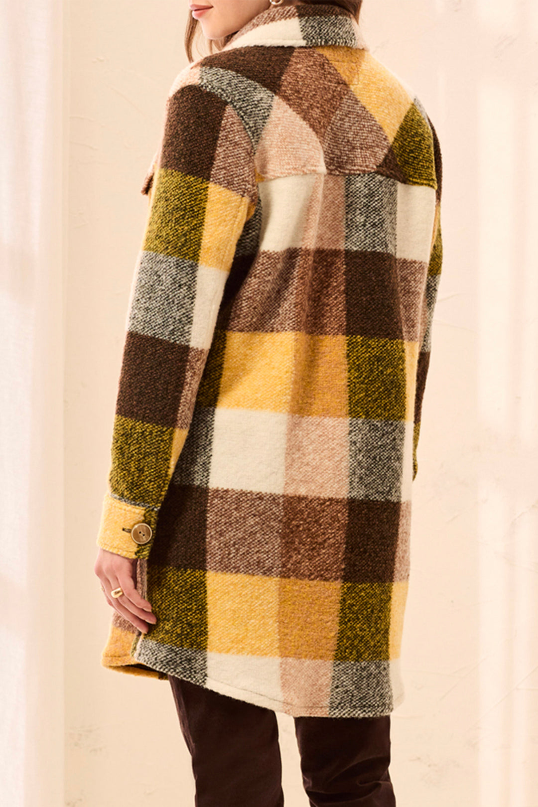 A woman shown from the back wearing a long plaid coat in mustard yellow, brown, cream, and olive green, paired with a beige knit sweater. 