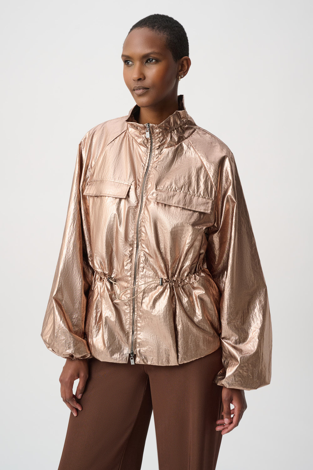 A model wearing a metallic rose gold lightweight jacket with a high collar, front zipper, and drawstring waist, paired with brown trousers, against a light gray studio background.