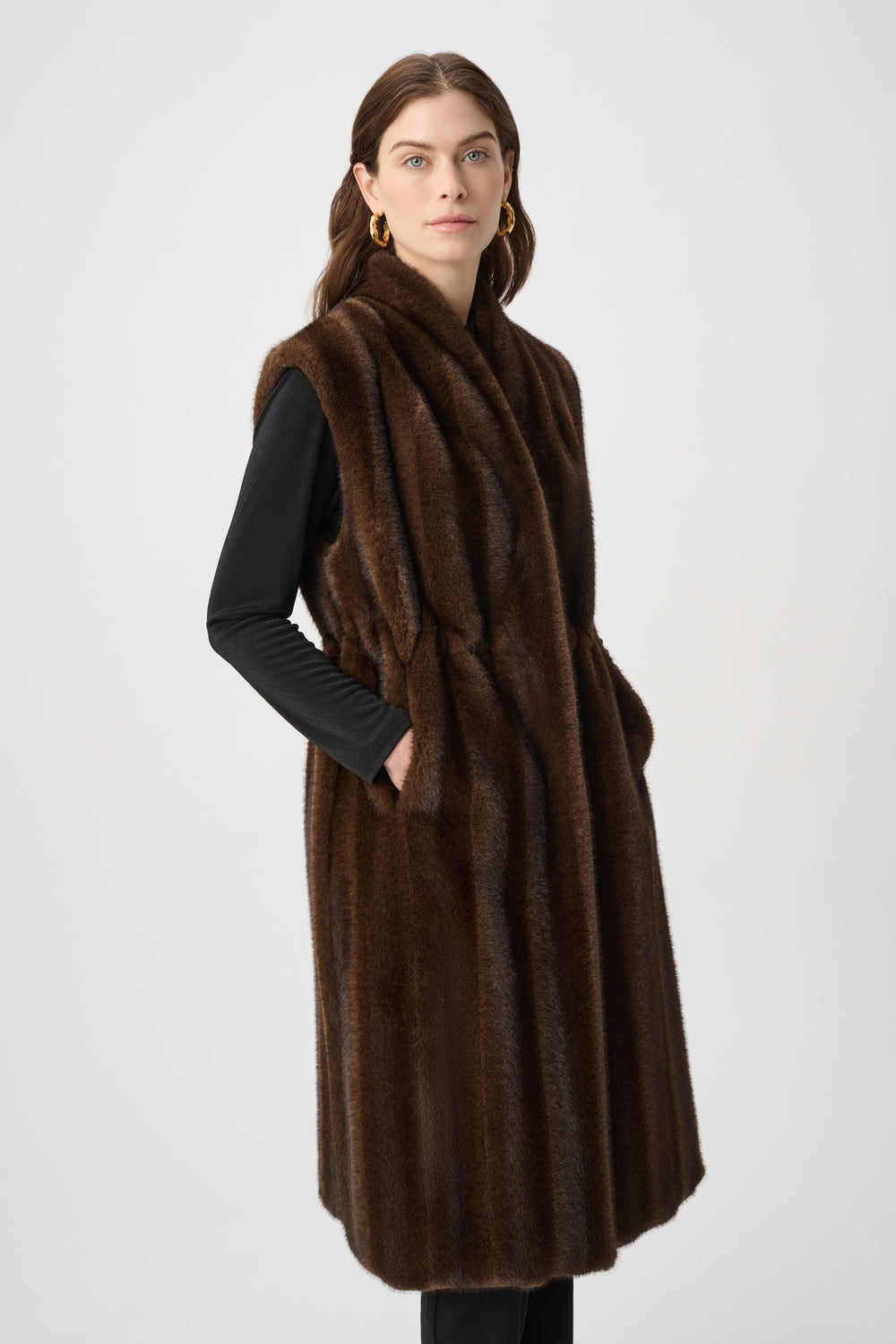 A woman standing in front of a light grey background wearing a long brown faux fur vest and black top and pants. 