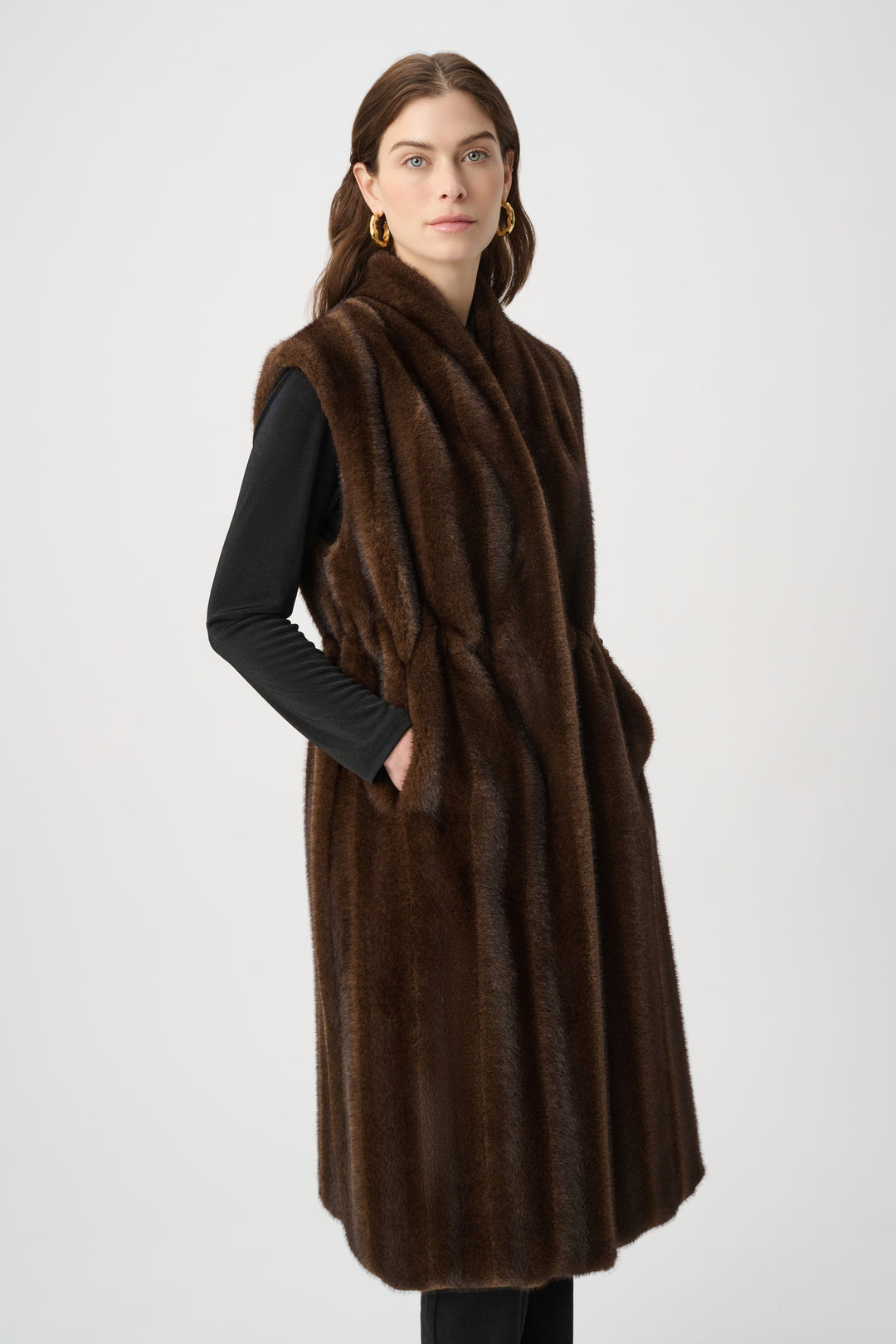 A woman standing in front of a light grey background wearing a long brown faux fur vest and black top and pants. 