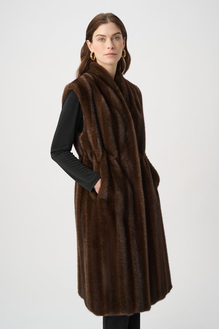 A woman standing in front of a light grey background wearing a long brown faux fur vest and black top and pants. 