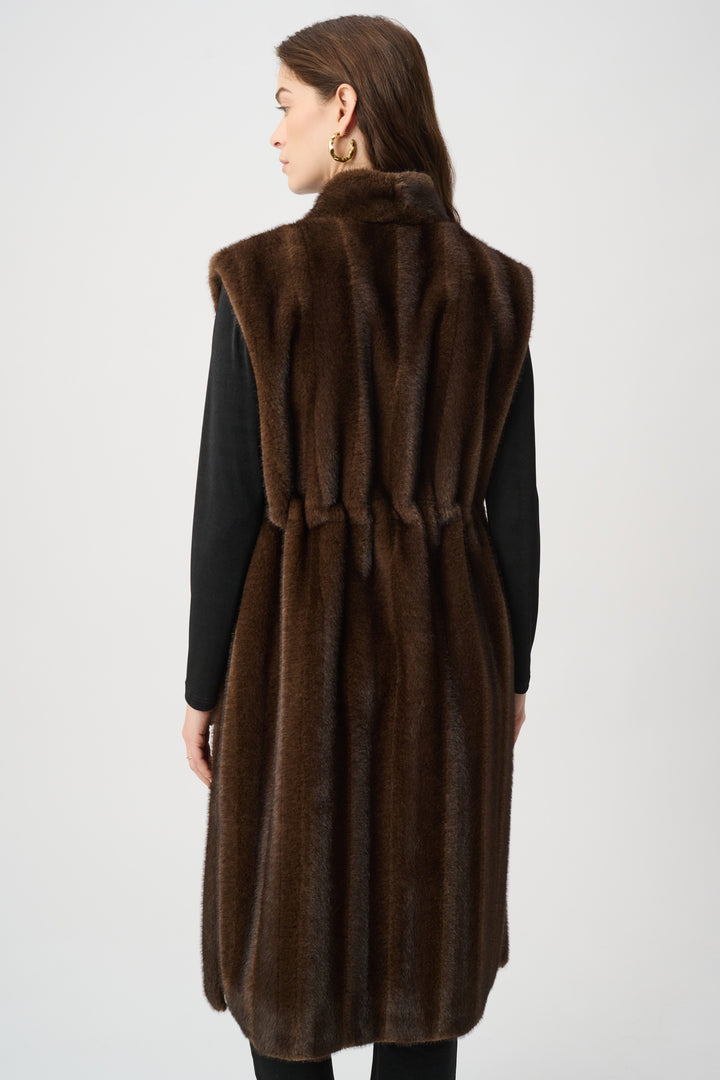 A woman shown from the back standing in front of a light grey background wearing a long brown faux fur vest and black top and pants. 