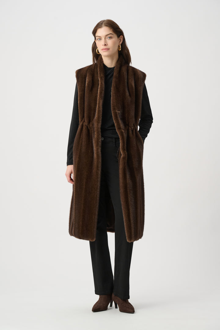 A woman standing in front of a light grey background wearing a long brown faux fur vest and black top and pants. 